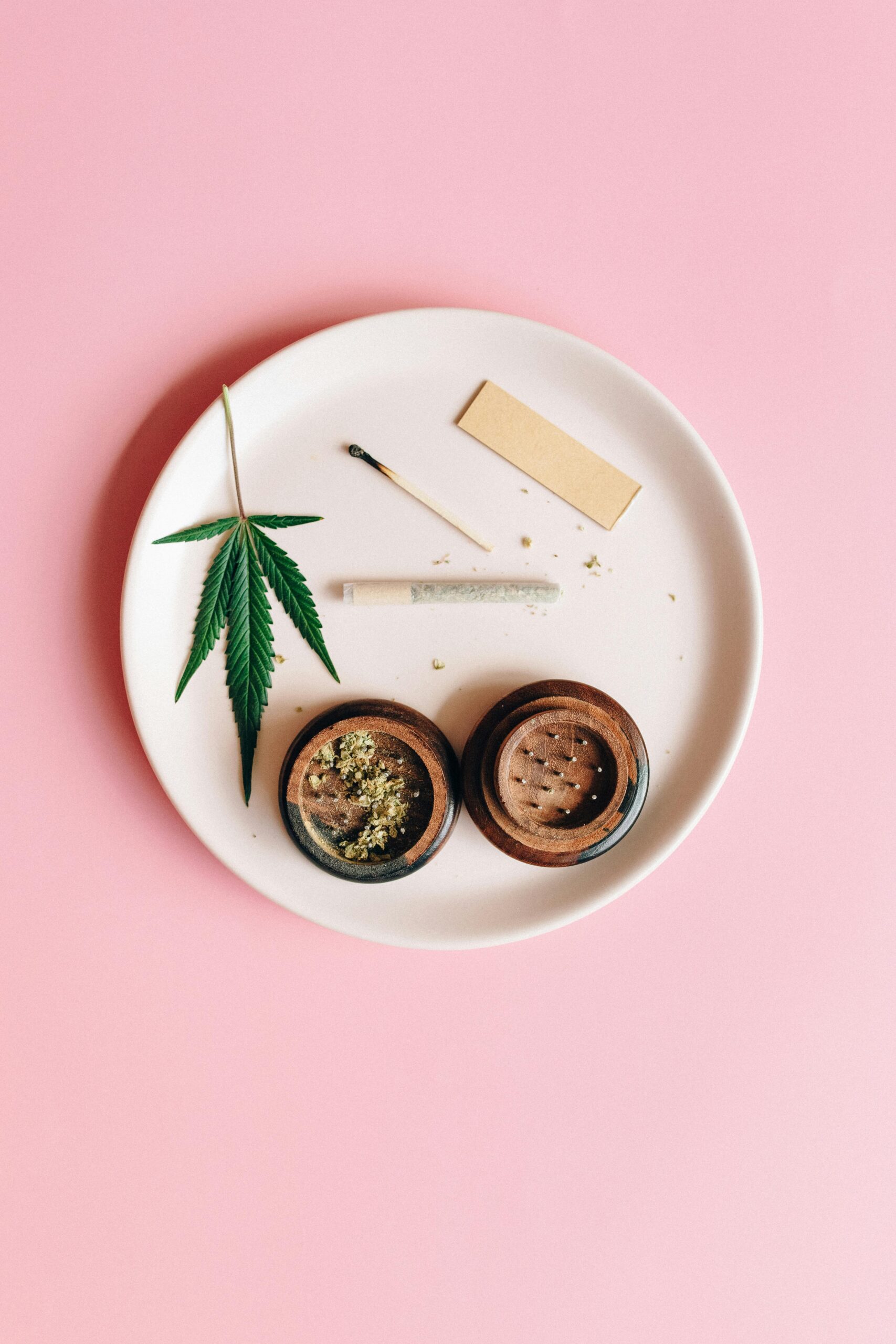 A cannabis leaf, grinder, and joint on a pink background, symbolizing relaxation.